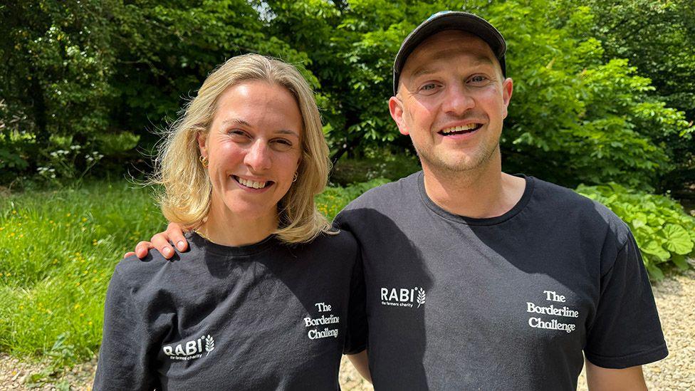 Siblings Alex and Hugh Addison, arm in arm and smiling. They're wearing dark blue t-shirts with the RABI charity logo and the words The Borderline Challenge. He is clean shaved and is wearing a baseball hat. She has shoulder-length blonde hair. Behind them are trees, bushes and grass.