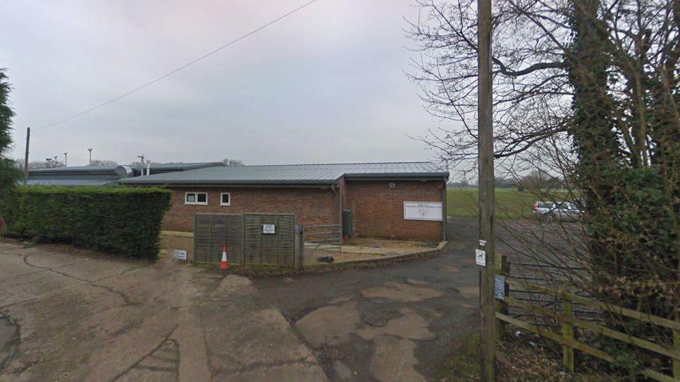 A single-story brick building with a metal roof at the end of a paved road. The building has a small fenced area in front, with a traffic cone placed near the gate. To the left, there is a tall hedge, and to the right, there are leafless trees and an open field with a rugby goal in the background. A sign is mounted on the building's wall, but its text is not legible.