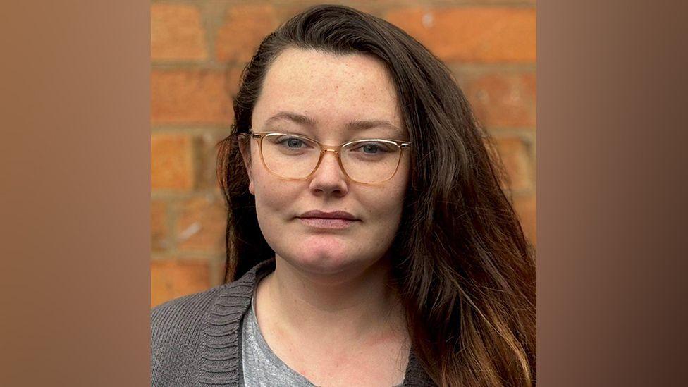 A headshot of a woman with long dark hair and glasses standing in front of a brick wall and looking at the camera.