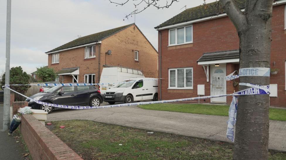 Police cordon around the front garden of a house, with vehicles in the drive, and police tape tied to a tree and lamppost