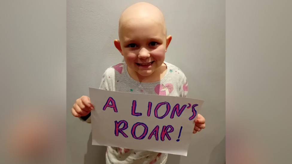 Lily holding a sign that says "a lion's roar" in pink and blue writing.