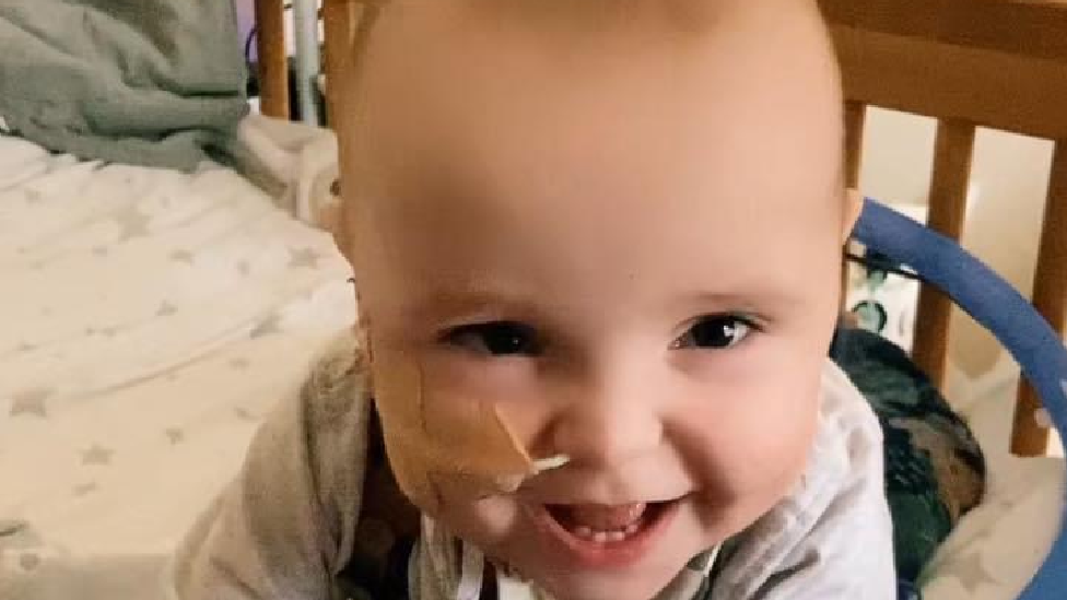 Oscar in his cot as a baby. He is looking at the camera smiling. He has a tube going into his nose. 