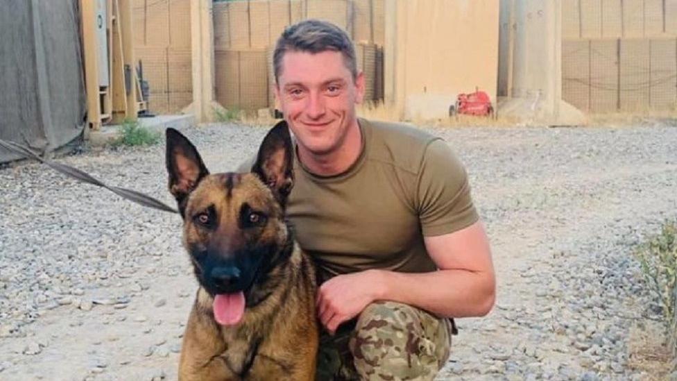 Nicki Hart in his army trousers and t-shirt, smiling next to a dog