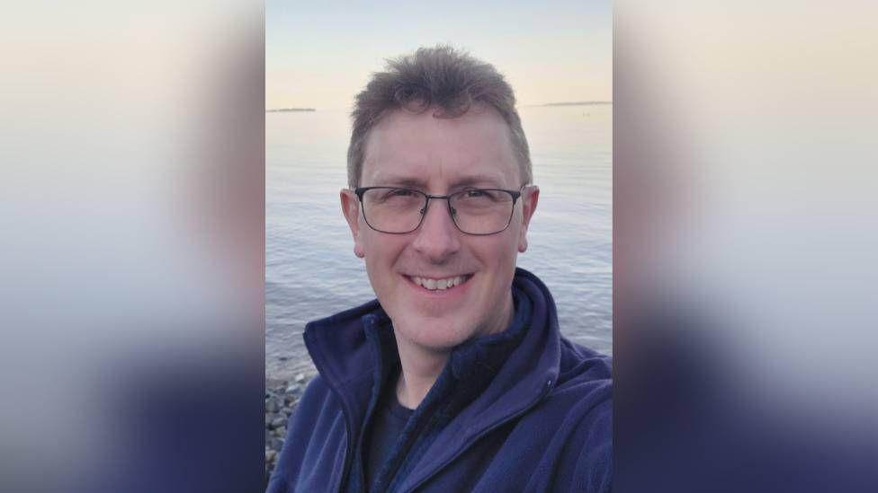 A selfie of Matthew Turner with a pebbled beach and sea behind him.