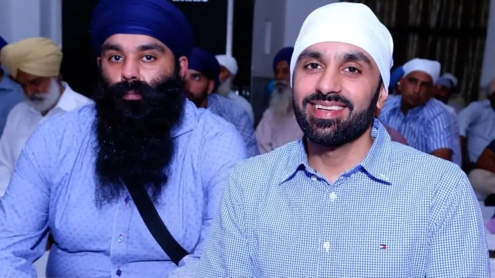 Gurpreet and Jagtar Singh Johal sit in a room with other Sikh men. Gurprret is wearing a royal blue turban and a light blue shirt and has a long, dark beard. Jagtar is smiling and wearing a white turban and a blue and white checked shirt.