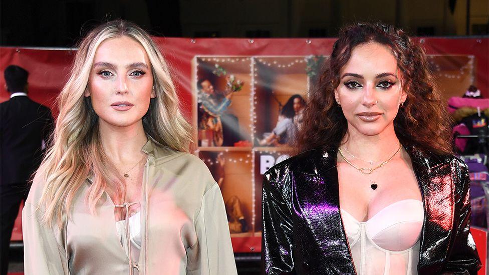 Perrie Edwards and Jade Thirlwall standing next to each other on a red carpet event in 2021. Perrie is wearing a cream coloured fleece, and Jade a black leather jacket.