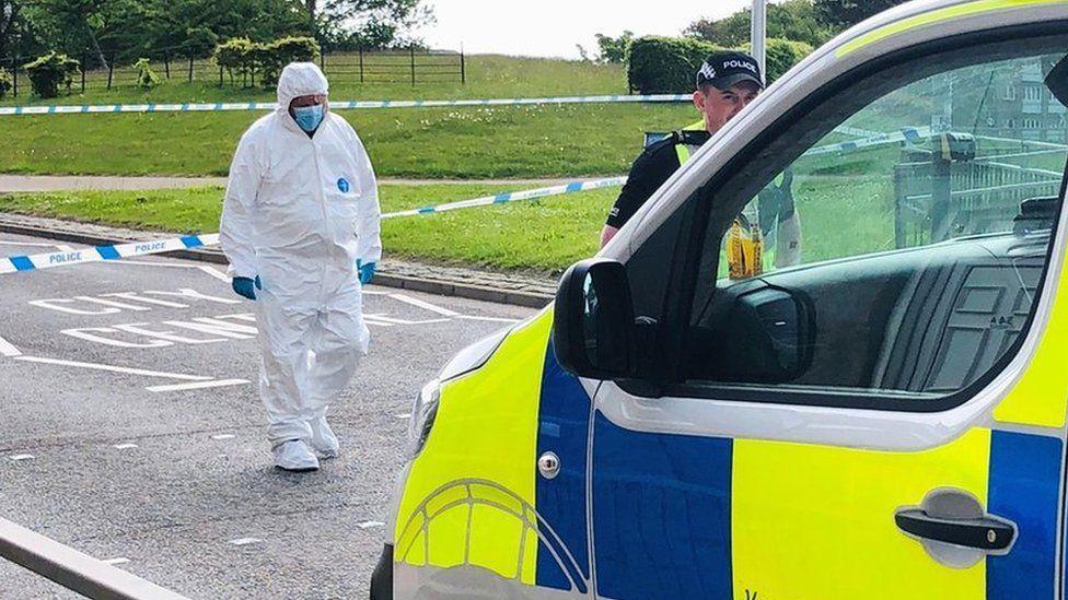 Forensic officer in white protective suit at scene of police incident, with police van in foreground