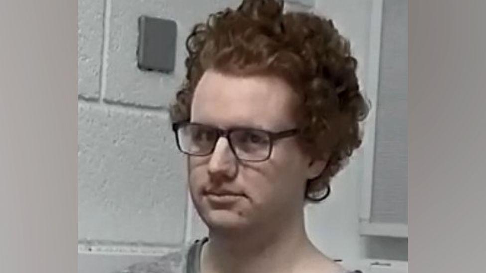 James Bubb has ginger hair and square glasses, standing by a white brick wall.