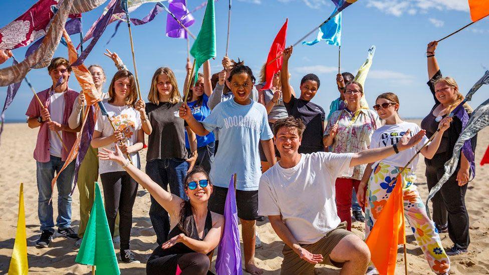 A group of young people on a sandy beach at Great Yarmouth.  They are wearing summery clothes and are waving colourful flags and looking happy. The sky behind them is blue.