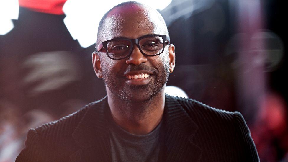 RaMell Ross at the Nickel Boys special screening during the 68th BFI London Film Festival at The Royal Festival Hall - he has black glasses on and is wearing a grey T-shirt and black corduroy jacket. He is smiling at the camera.