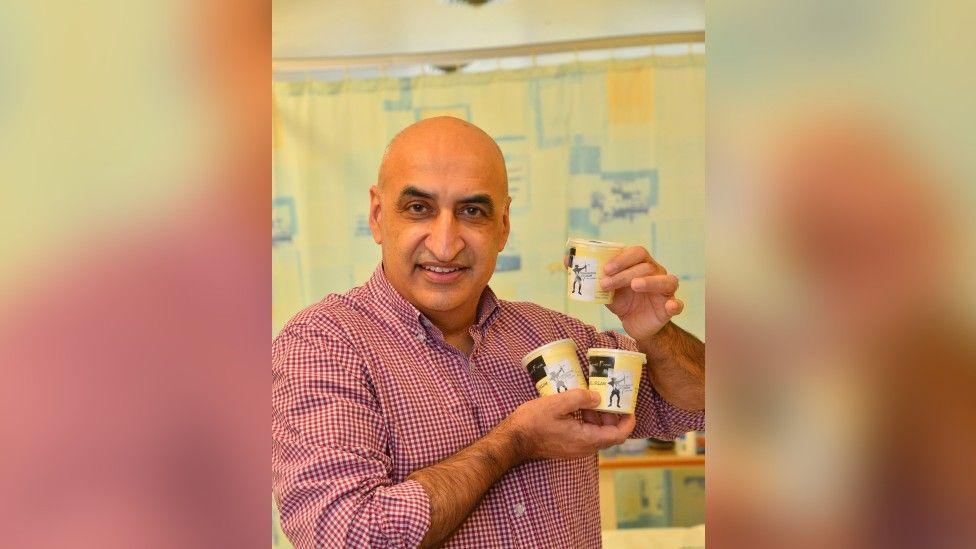 Professor Opinder Sahota stood holding some tubs of "N-ICE Cream", smiling at the camera.