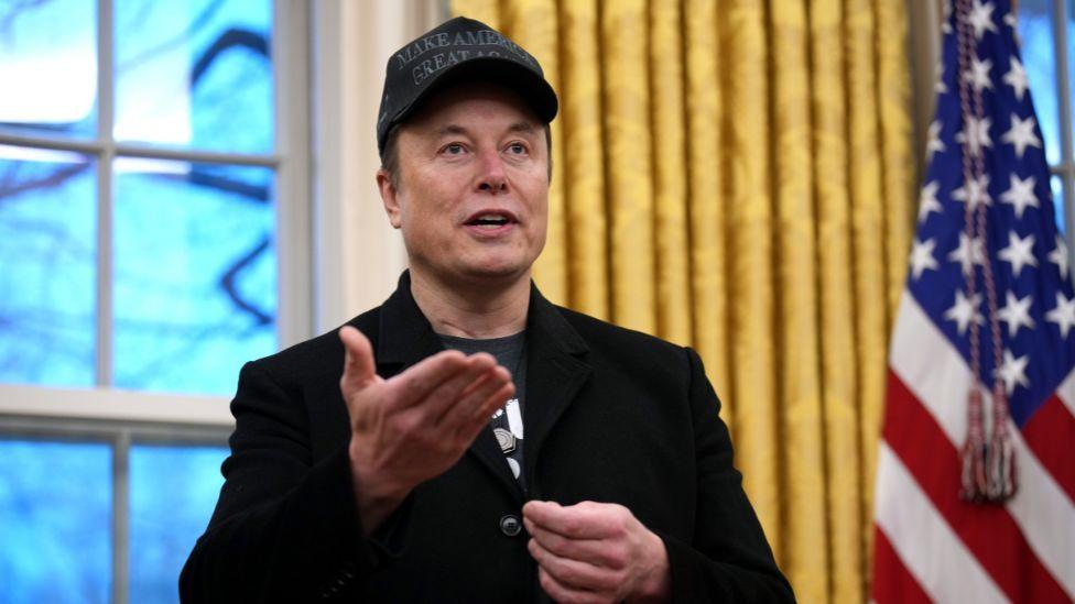 Elon Musk, a man with dark hair covered by a black "Make America Great Again" cap, speaks in the Oval Office. He is wearing all black, with his hands raised in front of him. He is visible from the chest up. Behind him are gold curtains, a window and a US flag. 