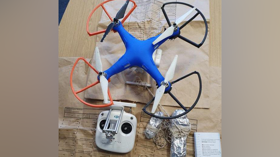 A blue drone with orange and black rotor shields and white blades on a wooden table with two tinfoil wraps and a string