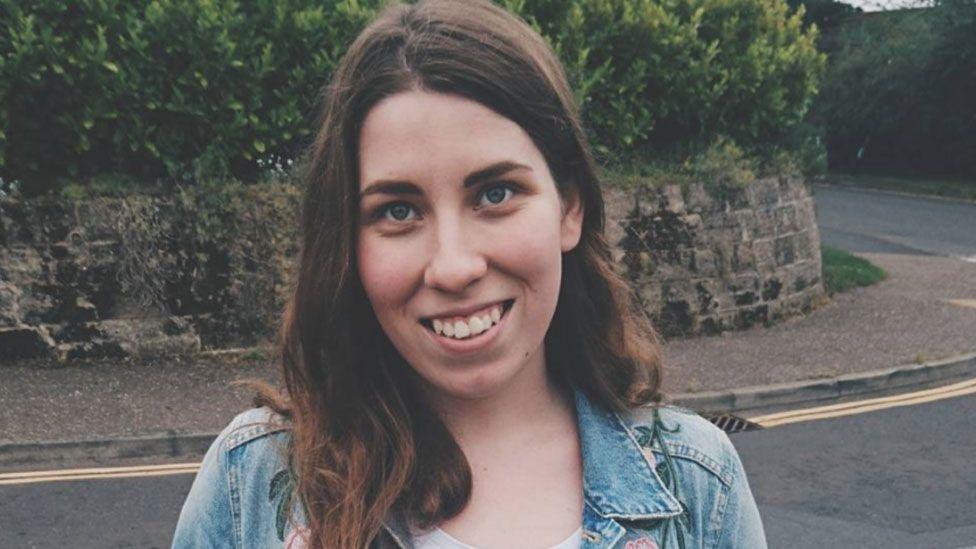 Sofia Price standing in a street with a low stone wall behind her. She has long dark hair swept over one shoulder and is wearing a light blue denim jacket over a just-glimpsed white top. She is smiling towards the camera