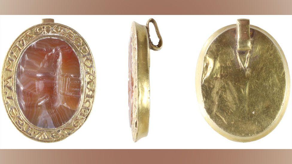 Three views of an oval medieval seal matrix, made from gold and a semi-precious gemstone. The view on the left shows its front. Its thin gold rim is etched with carved letters and surrounds the oval pink-toned gemstone, which is carved with an elephant with a castle or howdah on its back. The view in the middle is side on, showing a gold rim and a loop on its back. The view on the right shows plain gold back with a narrow gold rim and the loop at the top side. 