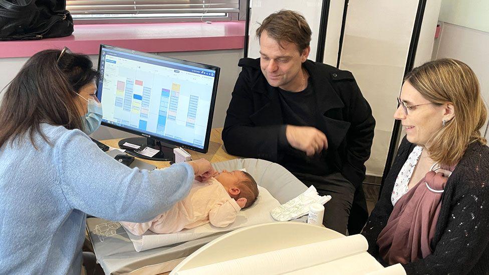 A baby wearing a light pink suit is weighed by a midwife with long brown hair and wearing a light blue jumper. The baby's parents, a brown-haired man and a blonde woman wearing glasses, look on and smile