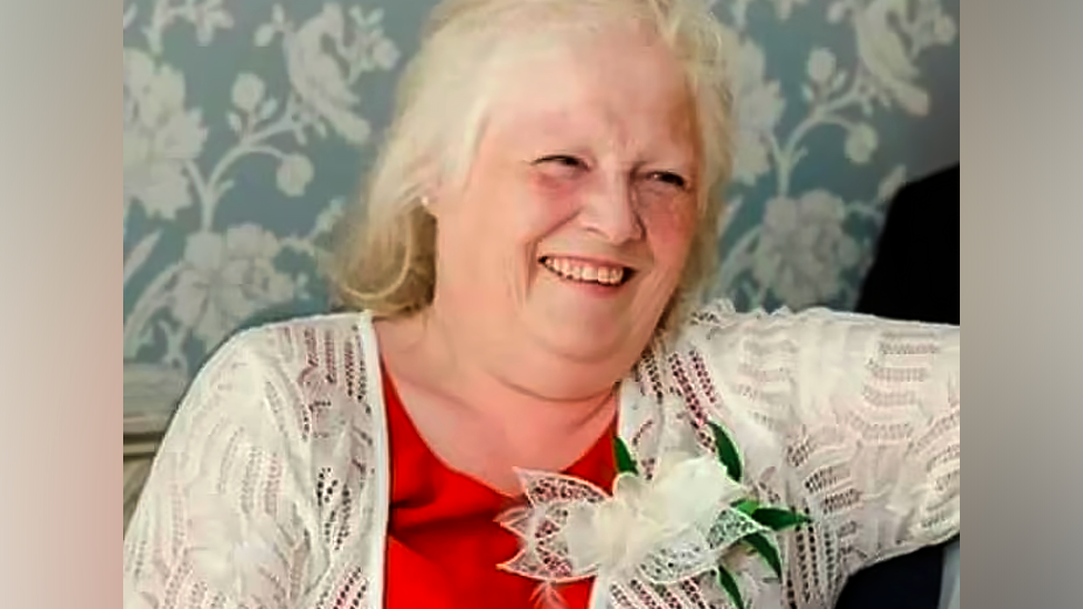 Esther Martin, who is wearing a red top and white cardigan with a flower pinned to it. She is smiling.