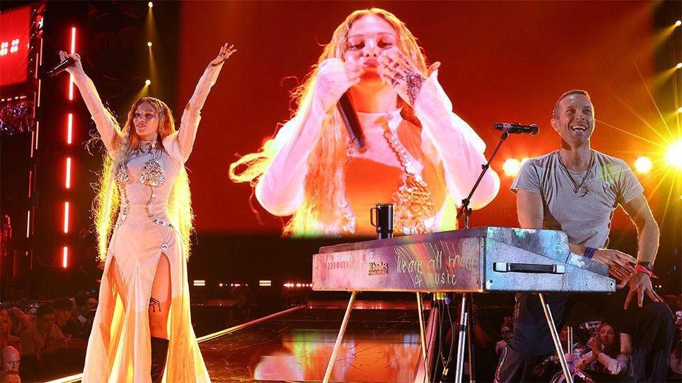 Elyanna and Chris Martin of Coldplay perform onstage at the 2024 iHeartRadio Music Festival in Las Vegas. Elyanna is wearing a long light coloured outfit and has her arms outstretched towards the sky. Her image is on the big screen behind her, while Chris Martin is sitting at a silver coloured keyboard, wearing a grey tshirt, smiling and looking out to the crowd.