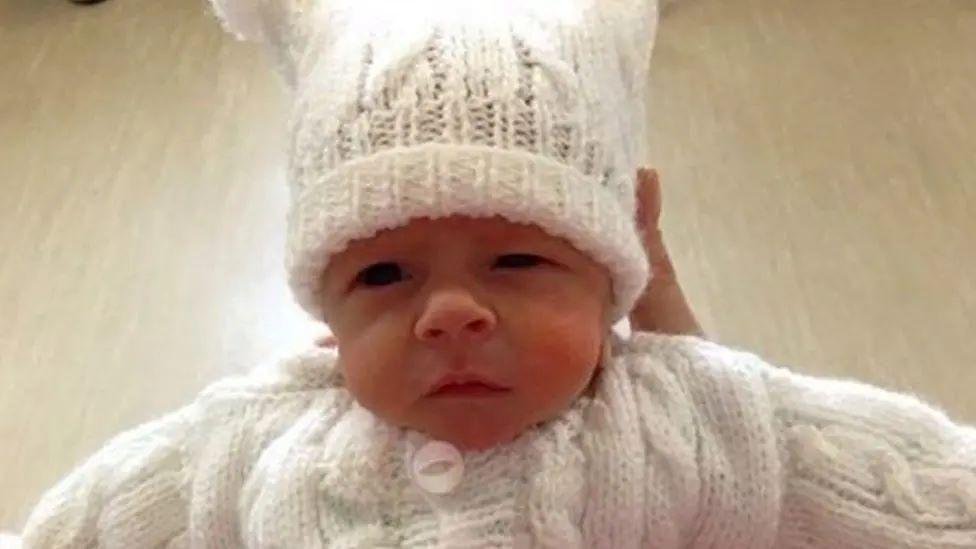 A new born baby wearing a white knitted hat and cardigan is held up to the camera. 