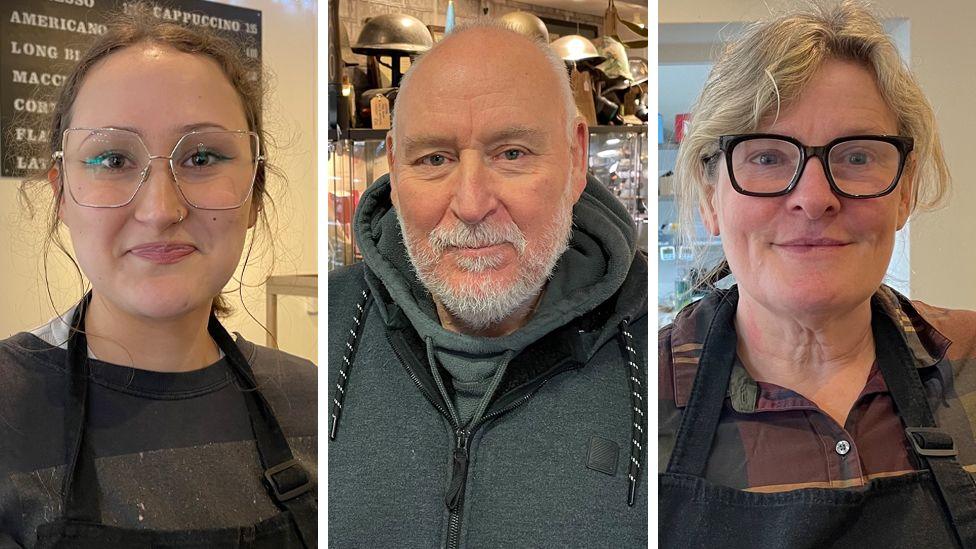 A composite image showing Lana Lacey, Lee Lawrence, and Lorraine Tidnam all looking down the camera. Lana is on the left of the shot. She is wearing large, square, wire-framed glasses over her brown eyes. She has brown hair that is pulled back away from her face and she is smiling a little. She is wearing a dark grey top and a black apron and is standing in front of a board on which are the names of various coffee-based drinks. In the middle of the picture is Lee Lawrence. He is wearing a dark green hooded top and has thinning grey hair, a full neatly trimmed grey beard and blue eyes. Behind him are soldiers' tin hats, mounted in a display. Lorraine Tidnam is on the right of the image. She is wearing rectangular glasses with thick black rims. Her blond hair is also worn up, away from her face. She is wearing a dark, checked shirt and a black apron.  