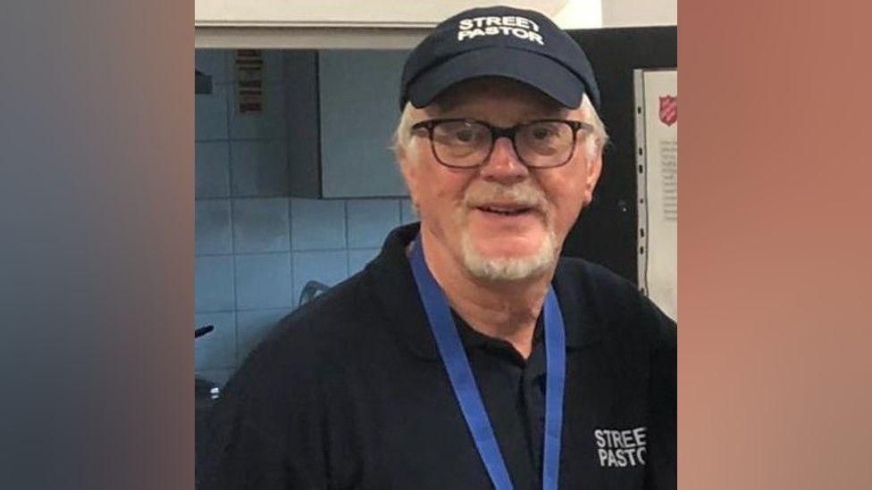 A man with white hair and a white beard and moustache wearing a black t-shirt that says £street pastor" on its breast. He has a blue lanyard around his neck, black glasses, and a black cap that says "street pastors".