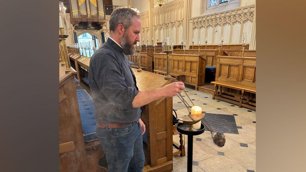 Matt Bullimore, chaplain of Corpus Christi College, Cambridge, with beard and short cropped greying hair, swinging a brass coloured thurible on brass-coloured chains through the air in the college chapel after setting light to the incense within