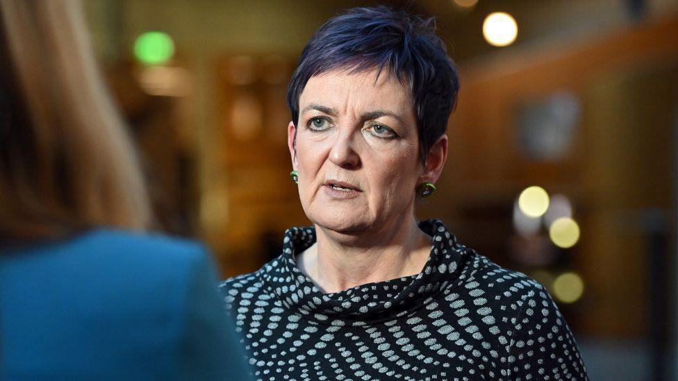 Angela Constance, who has short hair that is dyed purple, speaks to a  journalist in the Scottish Parliament. She is photographed over the shoulder of the journalist, and is wearing a black and green patterned top. 