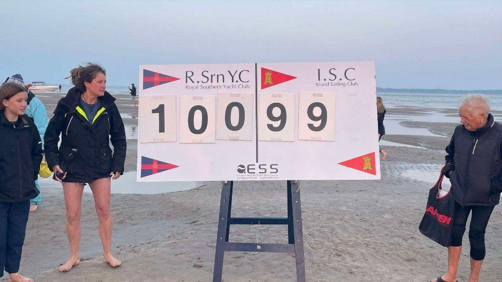 Cricket scoreboard on blue A-frame stands on sand with players either side