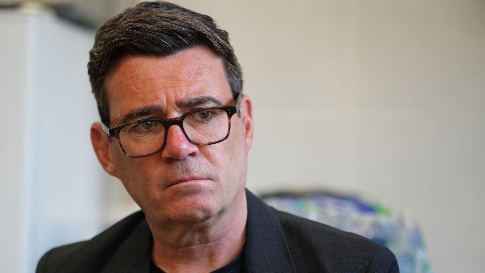 Andy Burnham, a man with dark hair and glasses. He is wearing a black suit jacket and t-shirt.