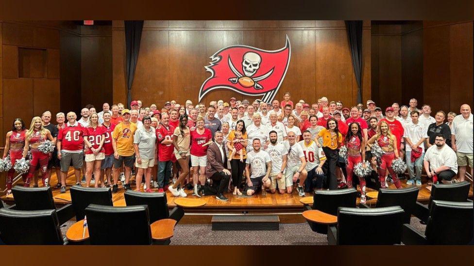 A large group of UK fans of the Tampa Bay Buccaneers stand in a group shot in a large room, under the team's flag, which is red with a large white skull and crossed swords on it. Many of the fans are wearing the team's red jersey.