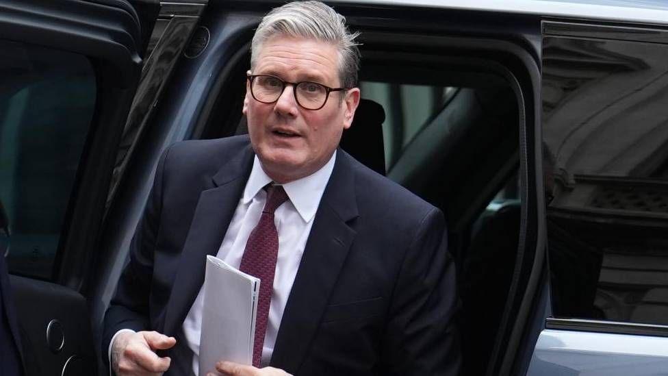 Sir Keir Starmer leaves from a vehicle with a piece of paper in his left hand. He has a neutral expression on his face.