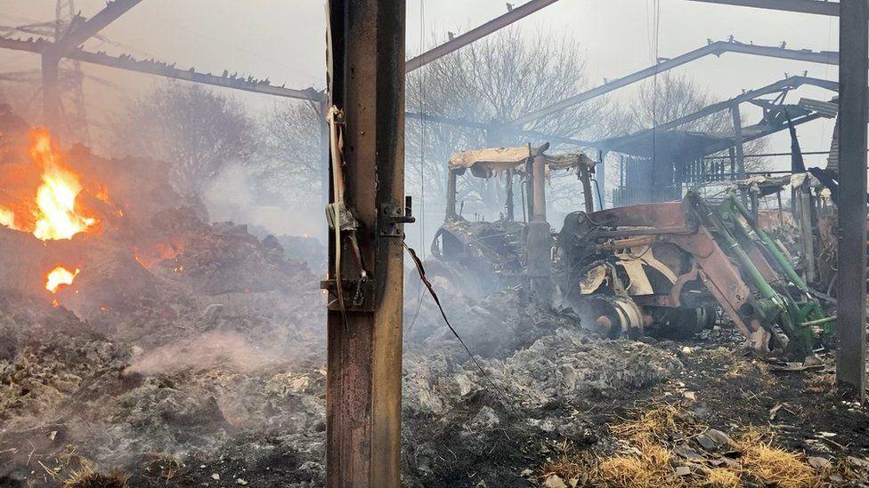 Charred shell of barn, flames can be seen in heap of material charred debris and several farm vehicles are burnt out in the barn.