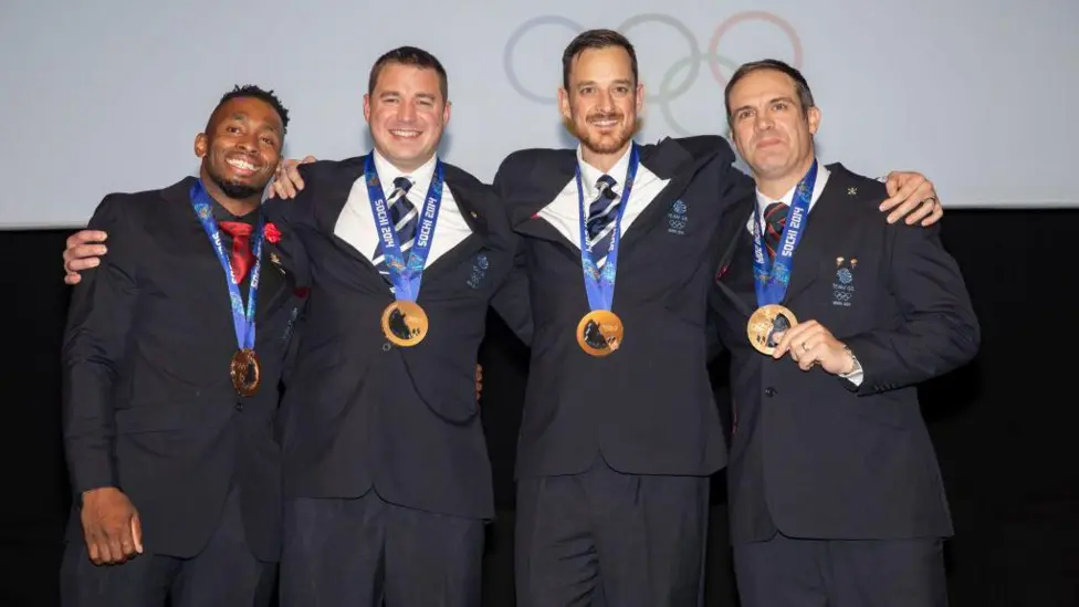Joel Fearon, Bruce Tasker, Stuart Benson and John Jackson, all wearing suits, stand with Winter Olympic bronze medals around their necks in 2019.