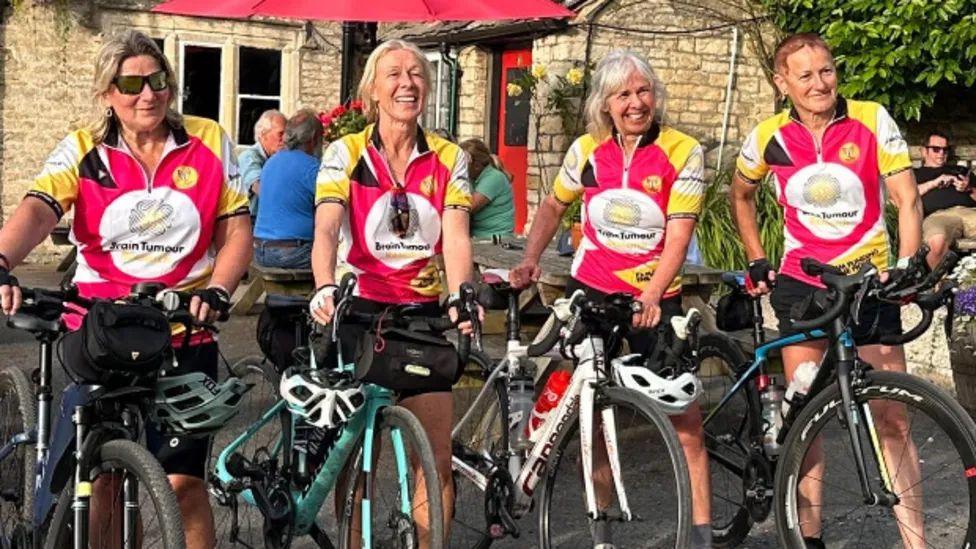 Four women stood with bikes. They are all wearing pink, white and yellow t-shirts with the Brain Tumour Research logo on the front. They are all smiling. 