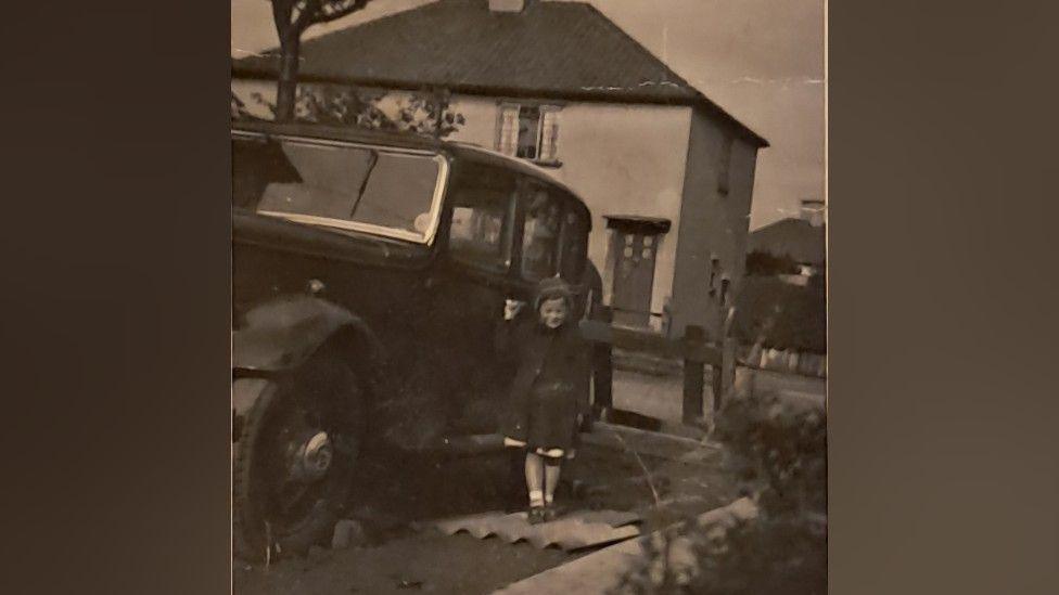 A black and white photo of a young girl holding onto the handle of a car door. The car is dark and large and old-fashioned. The girl is wearing a dress and a coat, with white socks and black shoes, and a hat on her head. There is a house in the background.