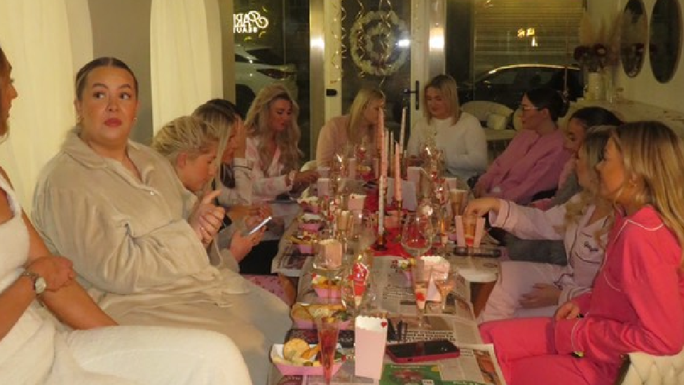 A group of women seated at a table, several in their pyjamas. The table has glasses, plates, and candles.