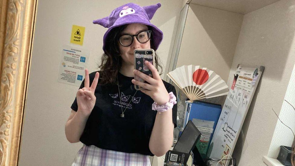 A person with shoulder-length wavy hair takes a selfie in a mirror, wearing a purple animal-shaped hat, a necklace and a black t-shirt. In the background is a computer and a fan with the Japanese flag