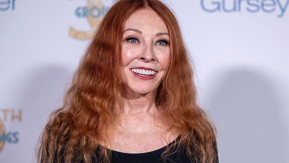 Cassandra Peterson, smiling at an event in California, wearing a black top and a white backdrop.