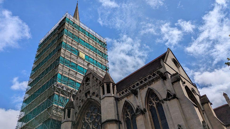 Exterior of the Church of Our Lady and the English Martyrs against a blue sky, with fluffy white clouds. The tower on the left is covered in scaffolding, while the dirty stone nave of the elaborate Victorian Gothic-style building is on the right.