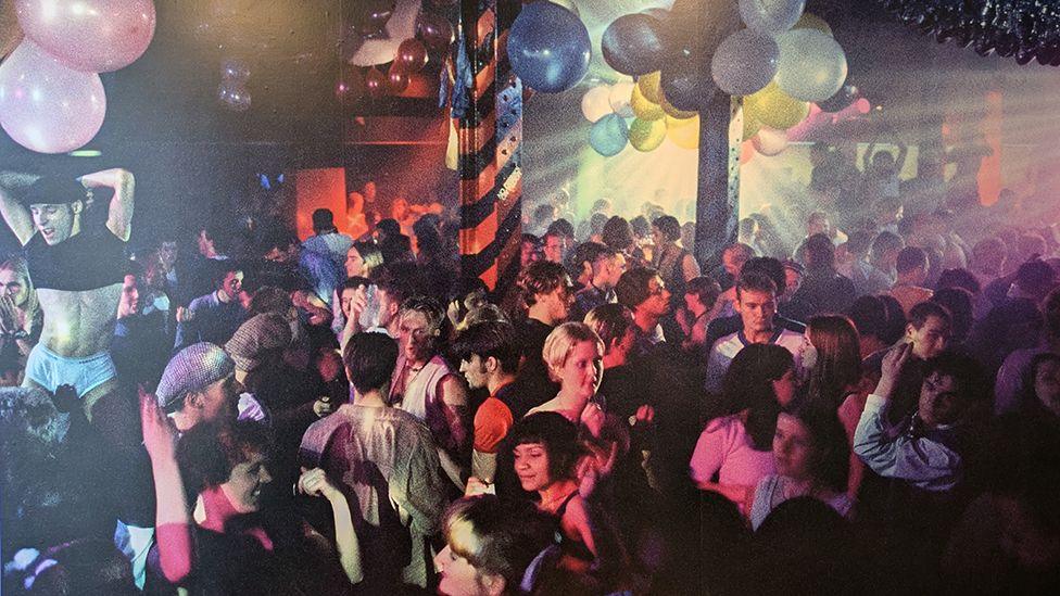 Dancers pack a club in the '90s with balloons grouped on the ceiling