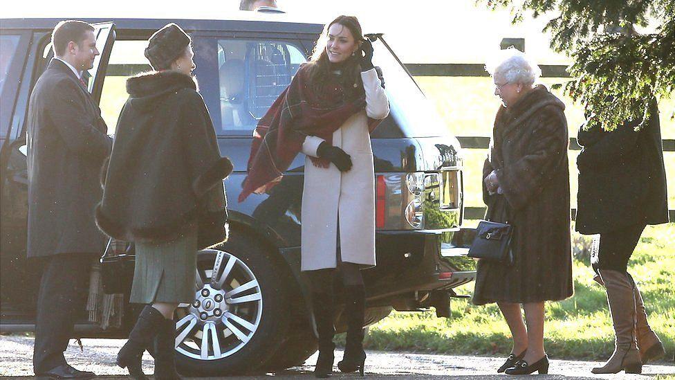 Princess of Wales stands in front of a car and is wearing a pale beige coat with a deep burgundy checked scarf. Princess Anne has a fur-lined cape and the late queen is wearing a dark brown fur coat. 