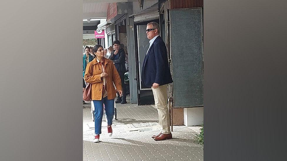 Hugh Bonneville standing outside Gadebridge Chemist in Hemel during filming of Paddington in Peru. He is seen sideways on wearing sunglasses, a blue jacket and cream chinos, and brown shoes. He is pouting and clenching his fists as though slightly irate. People are in the background looking on, and one lady wearing a tan coloured coat and blue jeans is walking towards him - she might also be a cast member. 