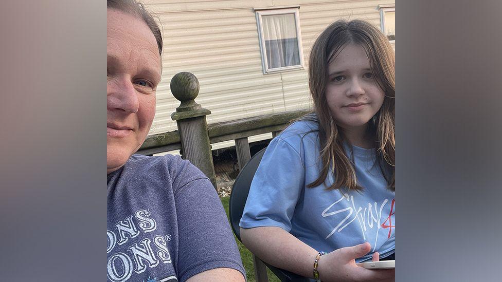 A selfie of Juile and Rosie. They are both sitting outside and there is a static caravan in the background. Julie is on the left of frame and only part of her face and body can be seen. She is smiling. Rosie is on the right of frame and is looking down the camera. She has brown, straight hair that is falling below her shoulders. Both Rosie and Julie are wearing T-shirts.