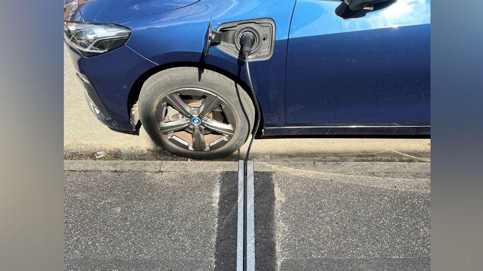 A close-up shot of a blue car with an EV cable plugged into the front panel, which slots into a channel in the pavement. The channel has metal runners on both sides side and darker bitumen where the pavement was dug out.