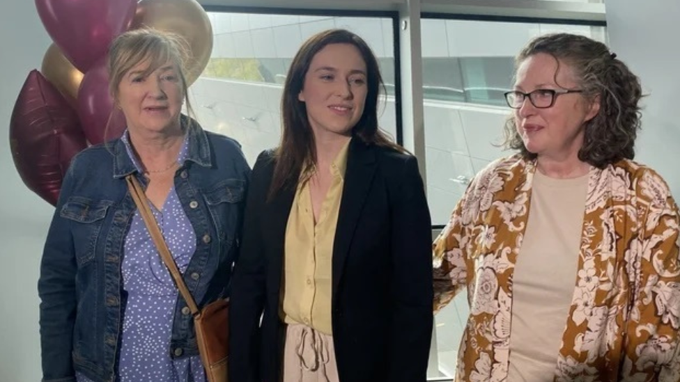 Tori Towey in a suit jacket with yellow blouse, smiling beside a collection of pink and gold balloons that say welcome home Tori, at Dublin Airport - her mother is in an orange patterned top and beige shirt, her aunt in a blue patterned dress and denim jacket with brown leather crossbody bag