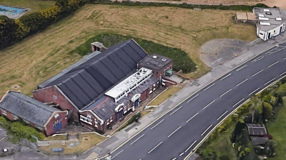 An aerial photograph of a rectangular building on edge of a road, set in a large green space.