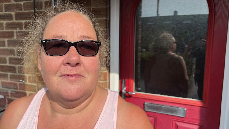 Lynda Magson outside her home in York. She is wearing a pink top and sunglasses and stood in front of a red door