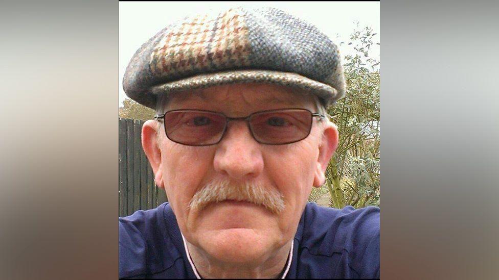 Keith Whiting wearing a hat and glasses. He has a moustache and is wearing a navy blue top