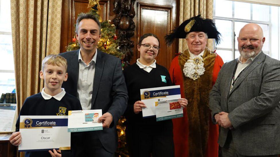 Oscar and Lilah at Hull Guildhall in their school uniforms holding their winners' certificates standing alongside the Lord Mayor in red robes and black feathered hat, leader Mike Ross and the council's chief executive  Matt Jukes who are wearing dark suits