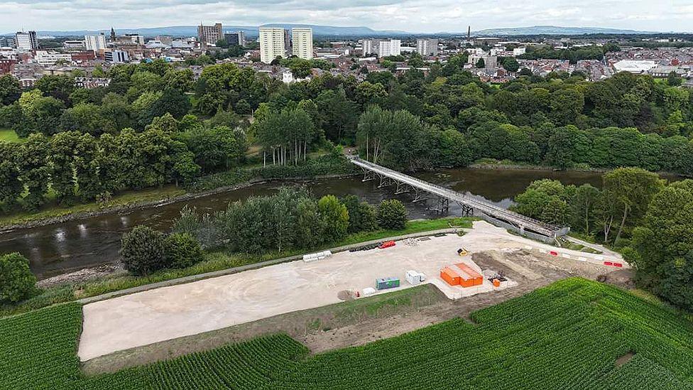 Aerial view of the site showing the river with the bridge across it, a large, flat area in the foreground with construction vehicles and metal storage units, and Preston city centre in the background